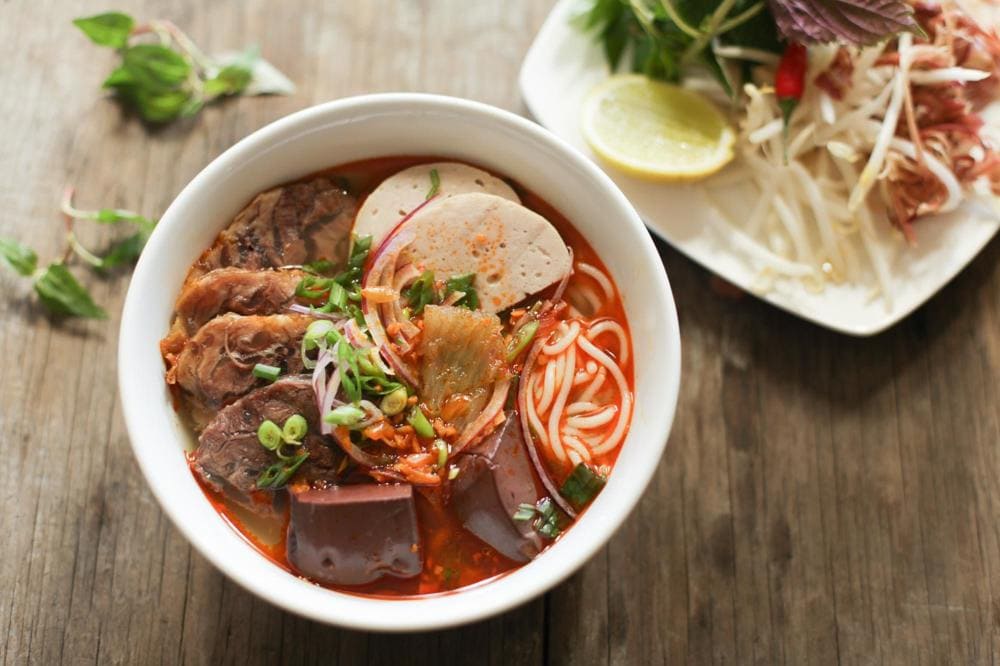 Vibrant red spicy noodle soup with thick rice noodles and herb garnishes